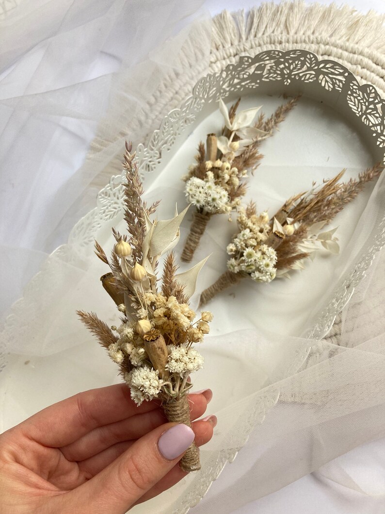 May include: Three small bouquets of dried flowers, tied with twine, in a white tray. The bouquets are made of white and beige dried flowers and grasses.