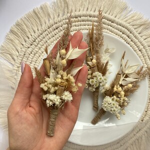 May include: Three small bouquets of dried flowers tied with twine. The bouquets are made with white and beige dried flowers and grasses. The bouquets are arranged on a white plate with a beige fringe around the edge.