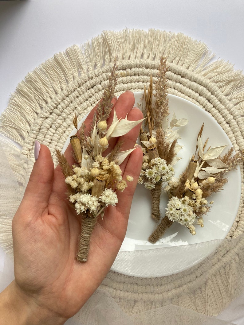 May include: Three small bouquets of dried flowers tied with twine. The bouquets are made with white and beige dried flowers and grasses. The bouquets are arranged on a white plate with a beige fringe around the edge.