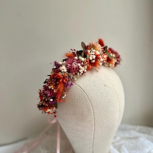 Può includere: Una corona di fiori secchi con fiori rosa, arancioni e bianchi. La corona è su una testa di manichino bianca e ha un nastro rosa legato intorno.
