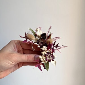 Könnte beinhalten: Ein kleiner Strauß getrockneter Blumen in den Farben Burgunderrot, Weiß und Grün. Der Strauß ist kreisförmig angeordnet und enthält zarte getrocknete Blumen und Gräser.