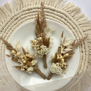 May include: Three small bouquets of dried flowers, each tied with twine, on a white plate. The bouquets are made of white and beige dried flowers and grasses. The plate is on a round, beige macrame placemat.