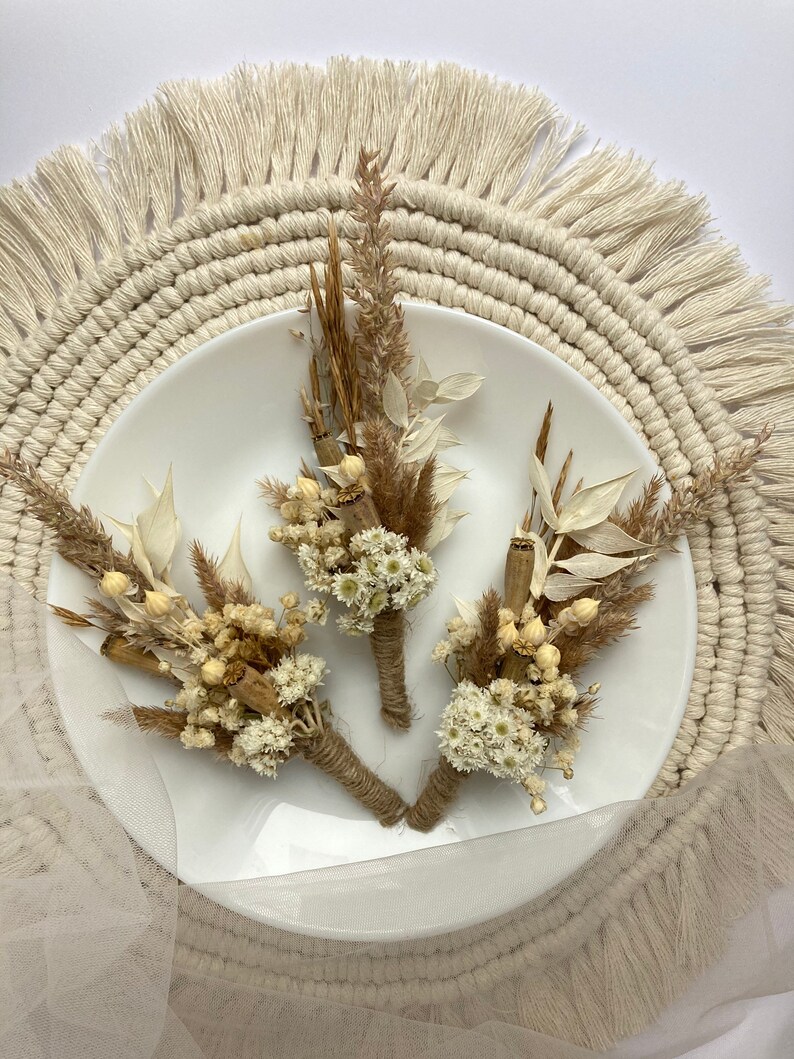 May include: Three small bouquets of dried flowers, each tied with twine, on a white plate. The bouquets are made of white and beige dried flowers and grasses. The plate is on a round, beige macrame placemat.
