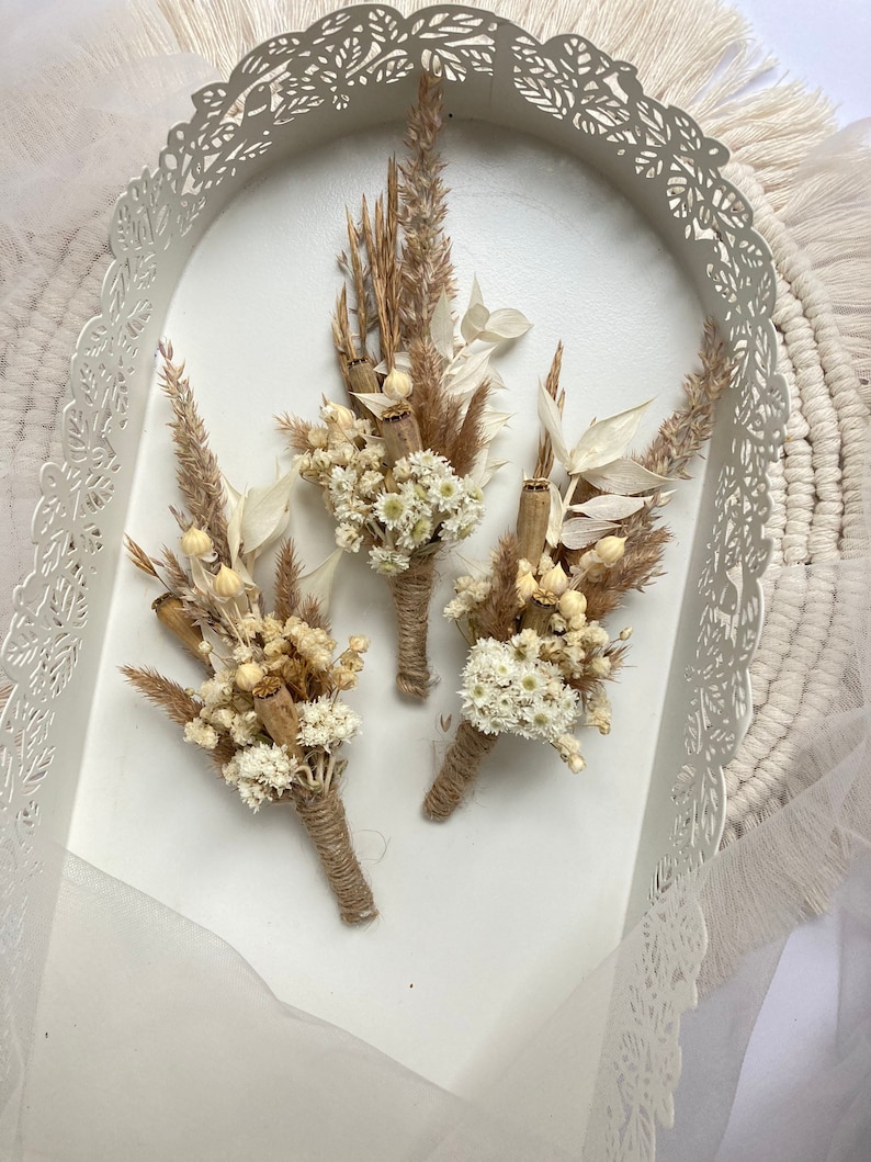 May include: Three small bouquets of dried flowers, each tied with twine. The bouquets are made with white and brown dried flowers and grasses. The bouquets are arranged in a white tray with a decorative cut-out design.