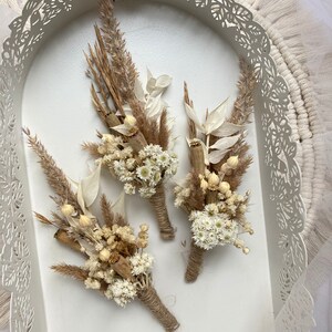 May include: Three small bouquets of dried flowers in shades of beige and white, tied with twine. The bouquets are arranged in a white tray with a decorative lace border.