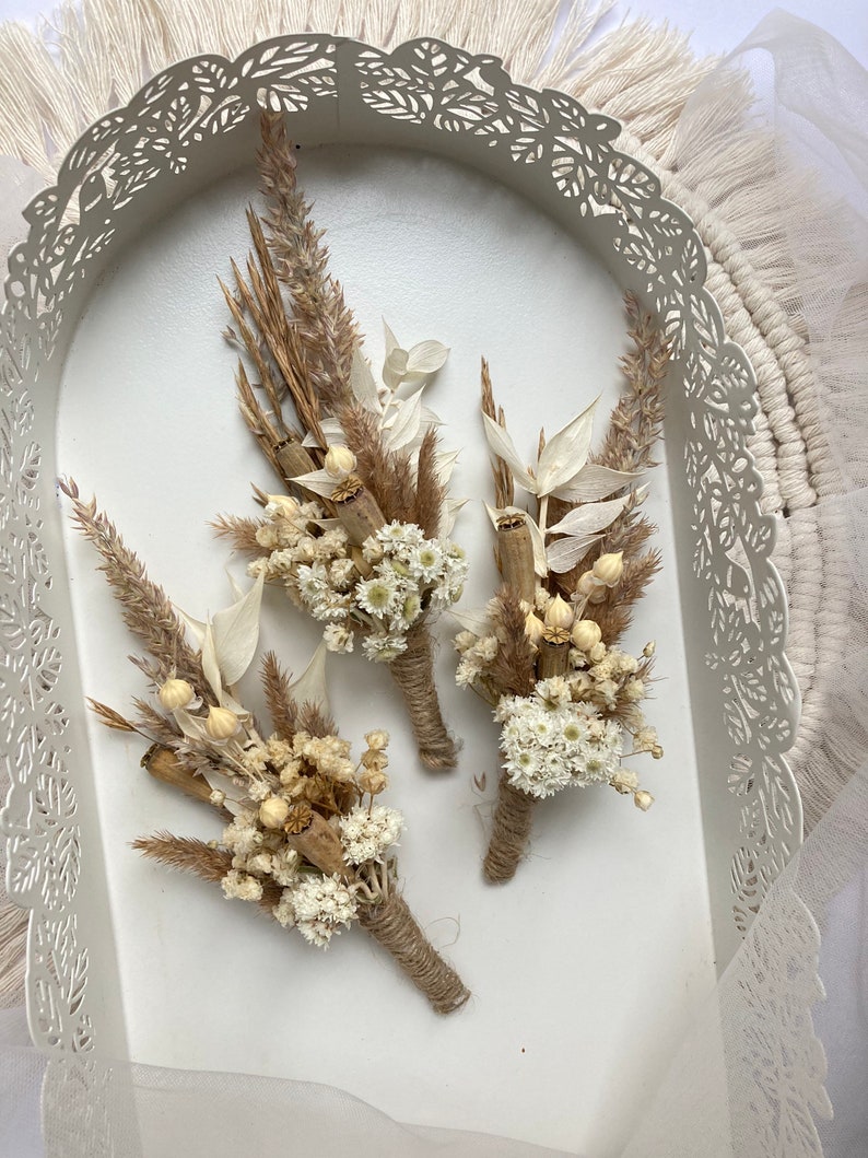 May include: Three small bouquets of dried flowers in shades of beige and white, tied with twine. The bouquets are arranged in a white tray with a decorative lace border.