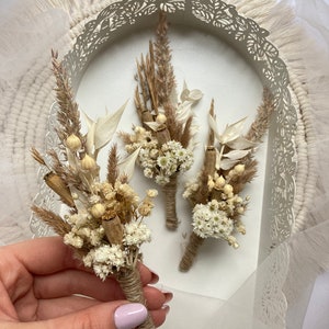 May include: Three small bouquets of dried flowers, tied with twine, in a white tray with a decorative edge. The bouquets are made with dried white flowers, brown grasses, and dried seed pods.