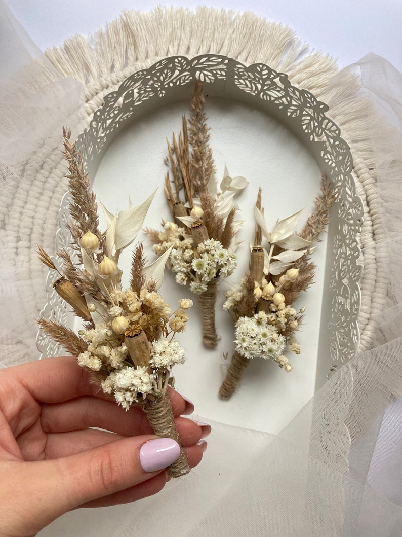 May include: Three small bouquets of dried flowers, tied with twine, in a white tray with a decorative edge. The bouquets are made with dried white flowers, brown grasses, and dried seed pods.
