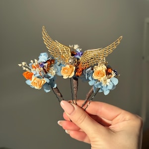 May include: Three gold hair pins with dried flowers. The flowers are blue, orange, and white. The top pin features a gold winged design.