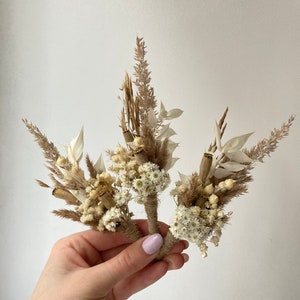 May include: Three small bouquets of dried flowers in shades of beige and white. The bouquets are tied with twine and feature dried grasses, seed pods, and small white flowers.