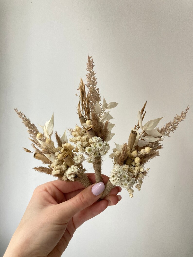 May include: Three small bouquets of dried flowers in shades of beige and white. The bouquets are tied with twine and feature dried grasses, seed pods, and small white flowers.