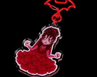 May include: A red and pink acrylic charm featuring a cartoon character wearing a red dress with a ruffled skirt. The charm is attached to a red plastic bat-shaped keychain.