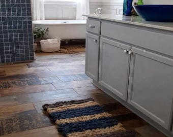May include: A rectangular, chunky knit bath mat featuring horizontal stripes in shades of dark blue, beige, and dark brown. The mat is placed on a floor with wood-look tiles in a bathroom setting that includes a gray vanity cabinet and a white tub.