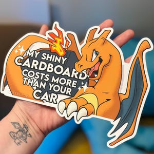 May include: A die-cut sticker featuring a cartoon Charizard in orange, yellow, and gray. The sticker has the text "MY SHINY CARDBOARD COSTS MORE THAN YOUR CAR". The sticker is held in a hand.