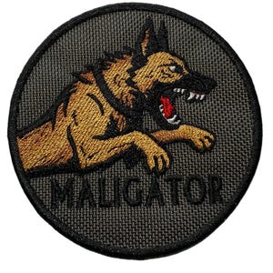 May include: Circular embroidered patch with a snarling German Shepherd dog in tan and black, set against a dark grey background. The dog's mouth is open, revealing teeth and a red interior. The word "MALIGATOR" is embroidered below.