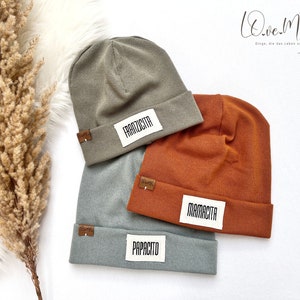 Könnte beinhalten: Drei Strickmützen in verschiedenen Farben mit den Worten "Franzicita", "Mamacita" und "Papaxito" darauf gedruckt. Die Mützen sind aus feinem Strickstoff gefertigt und sind ein beliebter Stil für Männer und Frauen.