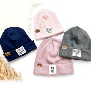 Peut inclure: Quatre bonnets en tricot de différentes couleurs avec un petit patch en cuir sur chacun. Les bonnets portent des phrases différentes : "Frech Dachs", "Zucker Suss", "Quatsch Kopf" et "Ähm No !"