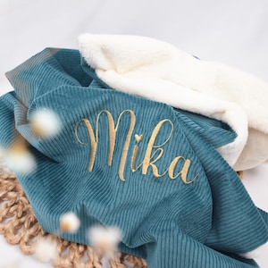 K&ouml;nnte beinhalten: Eine Babydecke aus tealfarbenem Cord mit dem Namen "Mika" in goldfarbenem Faden gestickt. Die Decke hat ein weiches, wei&szlig;es, pl&uuml;schiges Futter und ist &uuml;ber einer gewebten Matte gefaltet.