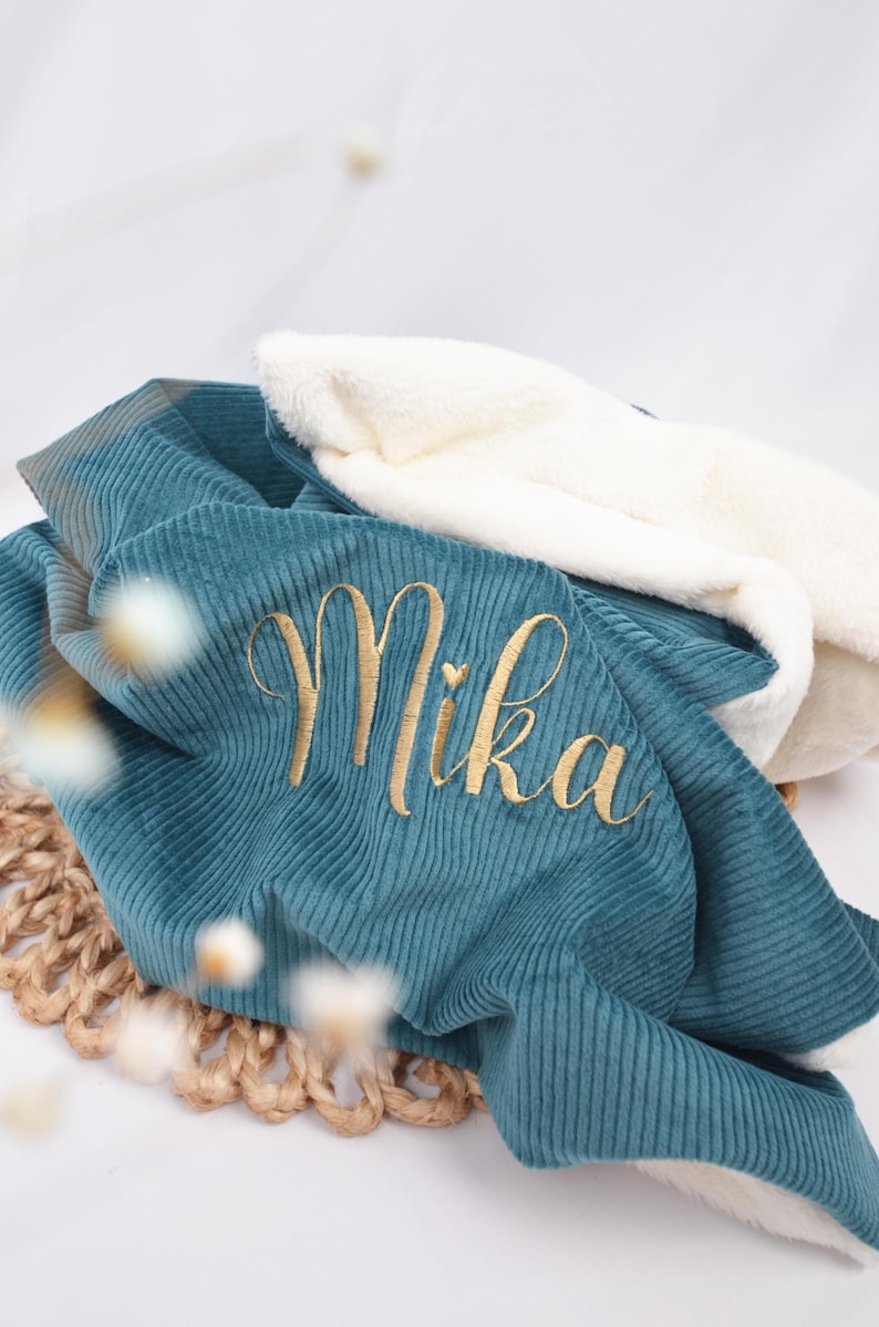 K&ouml;nnte beinhalten: Eine Babydecke aus tealfarbenem Cord mit dem Namen "Mika" in goldfarbenem Faden gestickt. Die Decke hat ein weiches, wei&szlig;es, pl&uuml;schiges Futter und ist &uuml;ber einer gewebten Matte gefaltet.