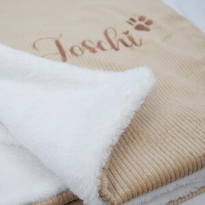 K&ouml;nnte beinhalten: Eine beigefarbene Corddecke mit wei&szlig;em, flauschigem Innenfutter. Auf der Decke ist der Name "Toschi" in Schreibschrift und ein Pfotenabdruck gestickt. Die Decke ist gefaltet und zeigt das weiche, wei&szlig;e Futter.