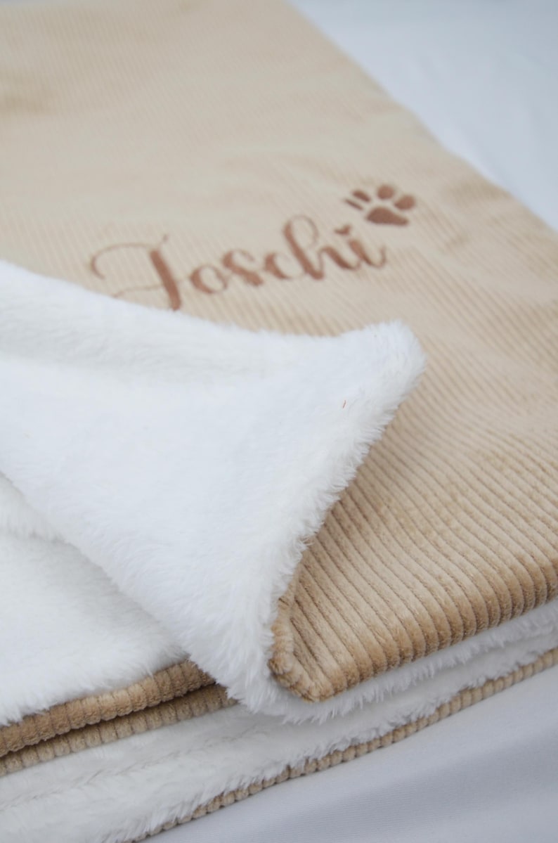 K&ouml;nnte beinhalten: Eine beigefarbene Corddecke mit wei&szlig;em, flauschigem Innenfutter. Auf der Decke ist der Name "Toschi" in Schreibschrift und ein Pfotenabdruck gestickt. Die Decke ist gefaltet und zeigt das weiche, wei&szlig;e Futter.