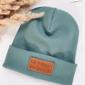 Könnte beinhalten: Eine tealblaue Mütze mit einem braunen Lederpatch, auf dem "Lieblingsmensch" in schwarzen Buchstaben steht.