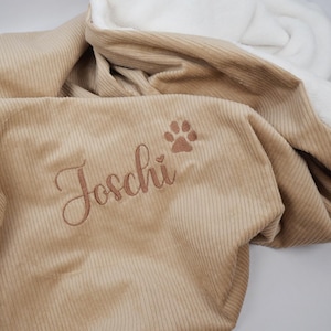 K&ouml;nnte beinhalten: Eine beige Corddecke mit wei&szlig;em, flauschigem Innenfutter. Die Decke hat den Namen "Joschi" in Schreibschrift gestickt, zusammen mit einem Pfotenabdruck. Die Decke ist gefaltet und zeigt beide Texturen.
