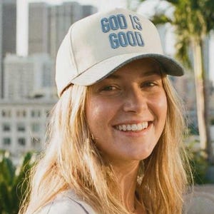 Könnte beinhalten: Beige Baseballkappe mit dem Text "GOD IS GOOD" in blauen Buchstaben. Die Kappe hat einen grauen Schirm. Der Hintergrund zeigt eine Stadtlandschaft und Palmen. Die Kappe wird von einer Person mit blonden Haaren getragen.