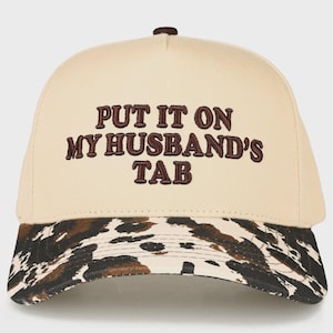May include: A beige baseball cap with the embroidered phrase "PUT IT ON MY HUSBAND'S TAB" in brown. The brim features a cowhide pattern in shades of brown and black. The cap has a brown button on top.