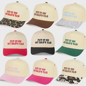 May include: A collection of cream-colored baseball caps with the text "PUT IT ON MY DAD'S TAB" embroidered in various colors. The brims are in different colors and patterns, including leopard print, brown, and black.