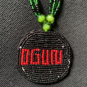Pu&ograve; includere: Una collana di perline nere con un ciondolo rotondo che presenta la parola "OGUDU" in perline rosse. La collana &egrave; realizzata con perline verdi e nere.
