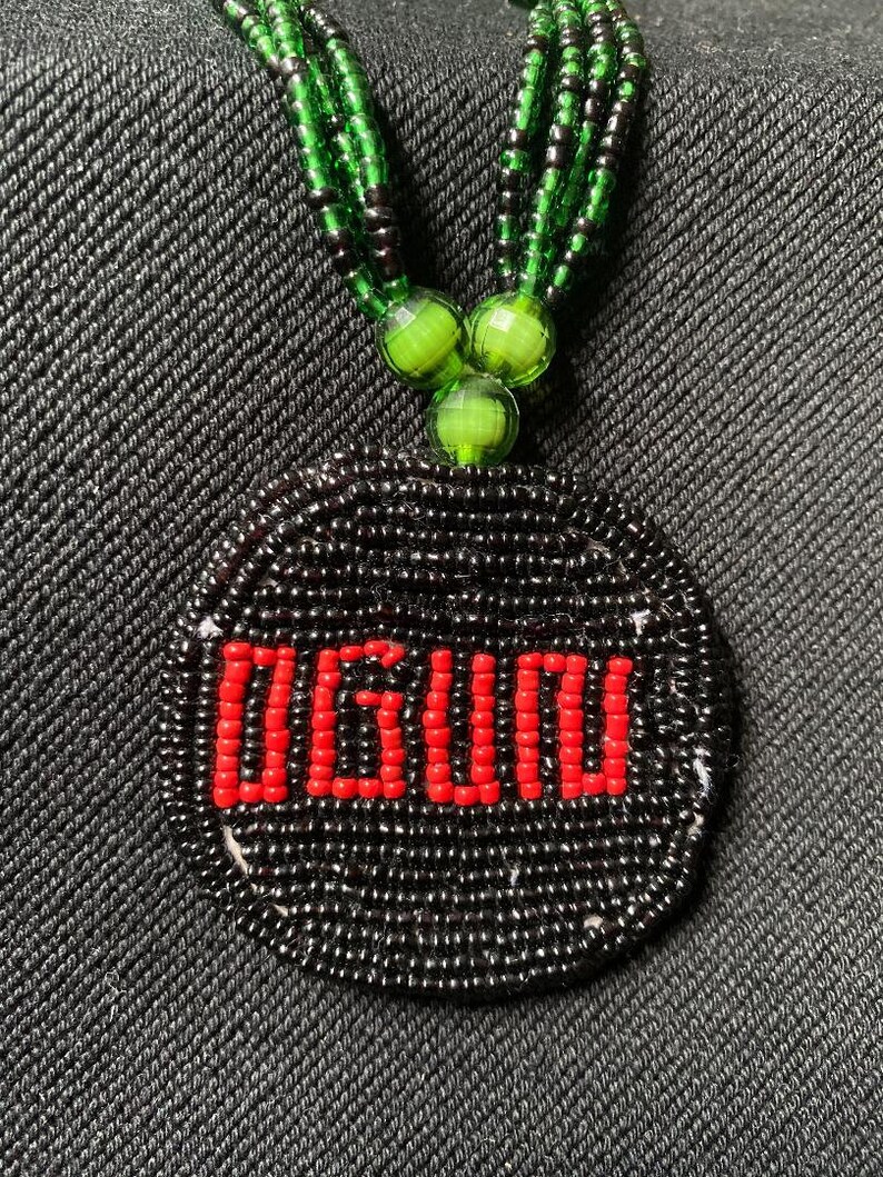 Pu&ograve; includere: Una collana di perline nere con un ciondolo rotondo che presenta la parola "OGUDU" in perline rosse. La collana &egrave; realizzata con perline verdi e nere.