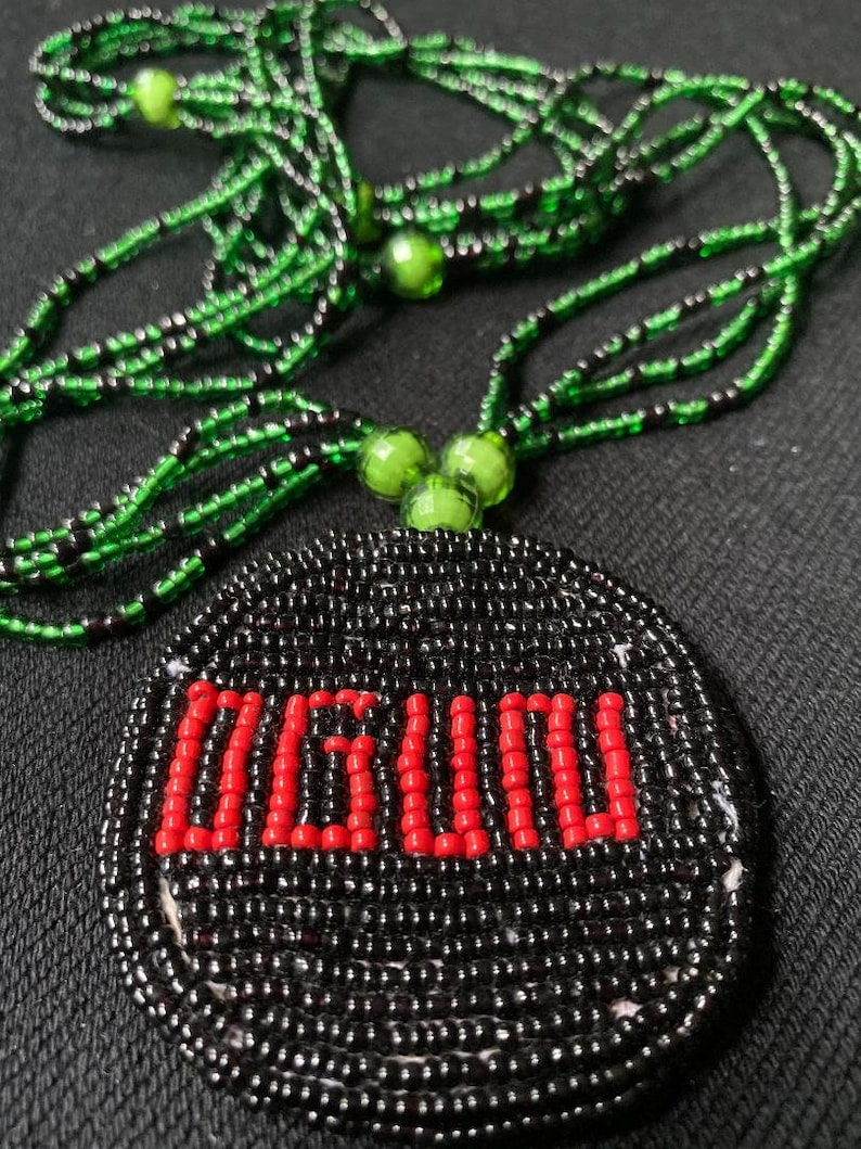 Pu&ograve; includere: Una collana di perline con un ciondolo rotondo che dice "OGUN" in perline rosse. La collana &egrave; fatta di perline nere e verdi.