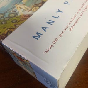 Pode incluir: Um livro branco com o t&iacute;tulo "The Story of Alchemy and the Philosopher's Stone" de Manly P. Hall. A capa do livro apresenta uma ilustra&ccedil;&atilde;o colorida de um mapa com pessoas e animais.