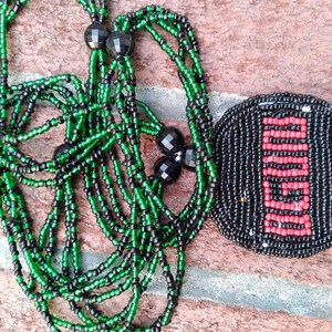 Pu&ograve; includere: Una collana di perline con perline verdi e nere, e un ciondolo rotondo di perline con un designo rosso e nero.