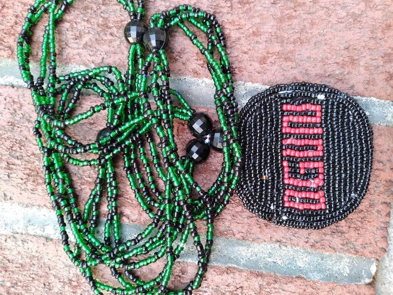 Pu&ograve; includere: Una collana di perline con perline verdi e nere, e un ciondolo rotondo di perline con un designo rosso e nero.