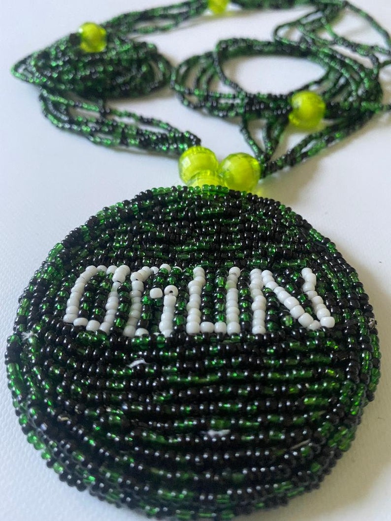 以下が含まれることがあります： 丸いペンダントが付いたビーズネックレス。ペンダントとネックレスは主に黒と緑で、「ORILU」の文字が白いビーズでペンダントに綴られています。ネックレスには緑色の球形のビーズが付いています。