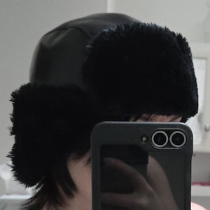 Può includere: Un cappello ushanka in finta pelliccia nera. Il cappello presenta una corona e paraorecchie in pelle nera con un'orlatura in finta pelliccia soffice. Il cappello è progettato per proteggere dal freddo.