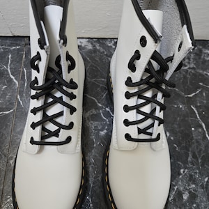 Puede incluir: Un par de botines blancos de cuero con cordones negros y costuras amarillas. Las botas tienen una punta redonda y una suela gruesa.