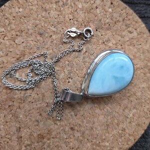 May include: A silver necklace with a teardrop pendant. The pendant has a light blue stone set in a silver bezel. The chain is a delicate silver link. The necklace is displayed on a brown surface.