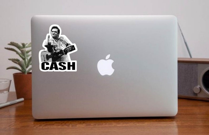 May include: A black and white sticker of Johnny Cash playing a guitar with the word "CASH" in bold letters below him.