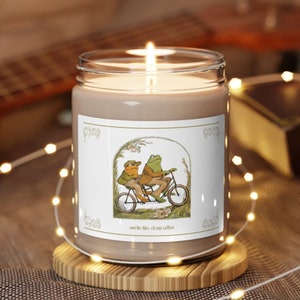 May include: A brown glass candle with a white label featuring a cartoon illustration of two frogs riding a bicycle. The label reads "smells like clean cotton".