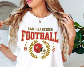 May include: White t-shirt with a red and gold graphic that says "San Francisco Football" with a football helmet and laurel wreath. The numbers 19 and 46 are on either side of the wreath.
