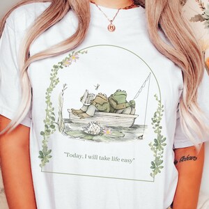 May include: White t-shirt with a green frog illustration. The frogs are in a boat on a pond with lily pads. The text "Today, I will take life easy" is printed below the illustration.