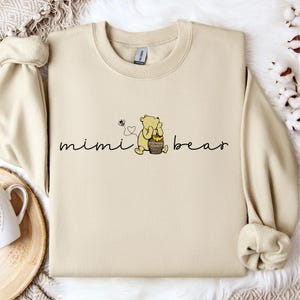 以下が含まれることがあります： ベージュのトレーナーに、くまのプーさんのイラストと「mimi bear」の文字がプリントされています。