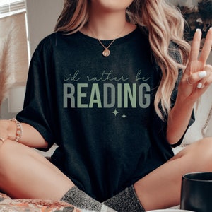 May include: A black t-shirt with the text "i'd rather be READING" in white and green lettering. The shirt is being worn by a person sitting on a bed.