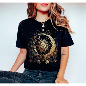 May include: A black t-shirt with a gold sun and moon design. The sun has a face and is surrounded by flowers and stars. The moon is crescent shaped and has a face on the side.