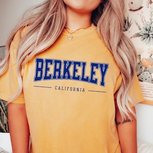 May include: Mustard yellow t-shirt with the word "BERKELEY" in navy blue block letters. Below the word is the word "CALIFORNIA" in navy blue, with a navy blue line above and below. The shirt is a casual style.