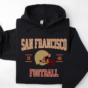 Puede incluir: Sudadera con capucha negra con un diseño gráfico dorado y rojo que dice "San Francisco Football" con un gráfico de casco de fútbol y los números 19 y 46.