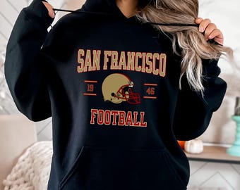 Op de afbeelding: Zwarte hoodie met een gouden en rode afbeelding met de tekst "San Francisco Football" met een afbeelding van een voetbalhelm in het midden.